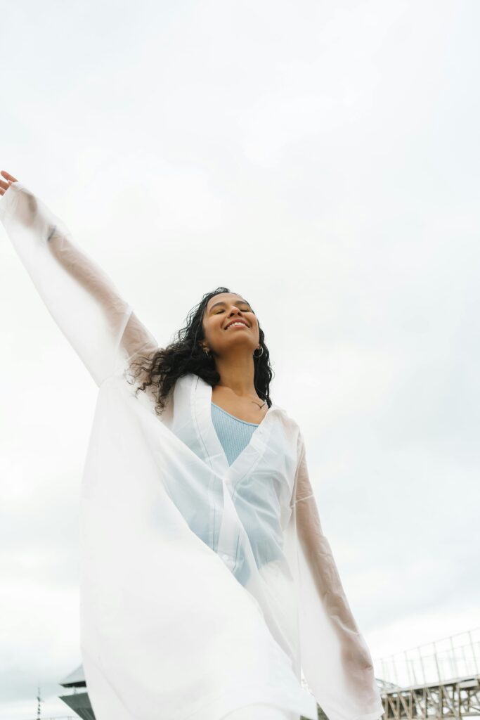 A joyful woman with curly hair wearing a white flowing garment enjoys a cloudy day outdoors.